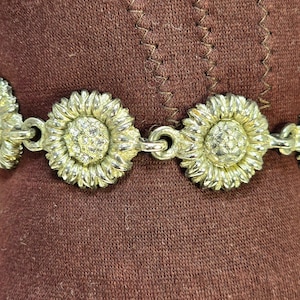 May include: A silver-toned bracelet featuring sunflower designs. Each sunflower has detailed petals and a center embellished with small, clear crystals. The bracelet is linked together with small, circular connectors. The background is a solid, dark brown fabric.