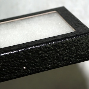 May include: Close-up of a black, textured box with a clear window and white interior padding. The box has a rectangular shape and a slightly glossy finish, suitable for jewelry or small items.