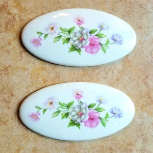 May include: Two white oval ceramic tiles with a floral design. The design features pink, white, and purple flowers with green leaves.