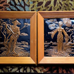 May include: Two framed wall art pieces. Each features a copper-colored relief of Asian figures in a landscape against a dark blue background. The frames are made of light brown wood.
