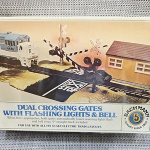 May include: A Bachmann Dual Crossing Gates model train accessory set in its original box. The packaging features a blue and white locomotive, a railroad crossing signal with black and white striped gates, and a small tan building.