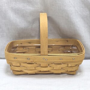 May include: A rectangular, light-colored woven basket with a tall, arched handle. The basket is made of thin, interwoven strips of wood, with a sturdy base and a clear plastic liner. The handle is attached with small metal rivets.