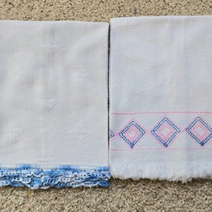 Puede incluir: Dos toallas de cocina blancas con detalles bordados. Una toalla tiene un borde de crochet azul y la otra tiene un patrón de diamantes rosa y azul.
