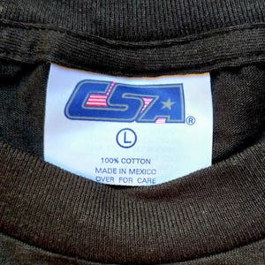 May include: Close-up of a black t-shirt collar with a white tag. The tag features the logo "CSA" in blue and pink, with a star and the letter "L" indicating size. Text on the tag reads "100% Cotton" and "Made in Mexico".