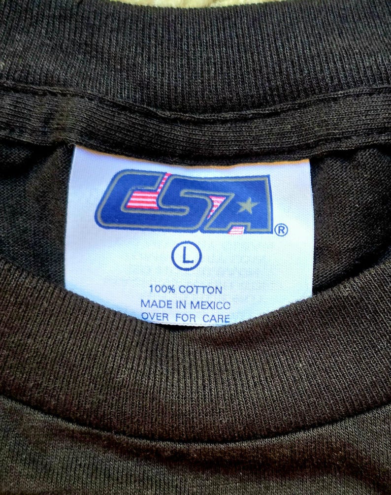 May include: Close-up of a black t-shirt collar with a white tag. The tag features the logo "CSA" in blue and pink, with a star and the letter "L" indicating size. Text on the tag reads "100% Cotton" and "Made in Mexico".