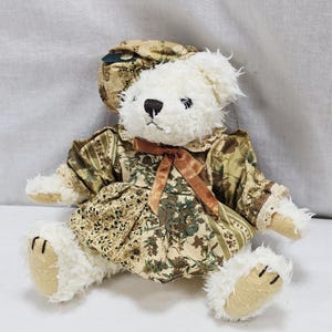 Op de afbeelding: Een witte teddybeer met een jurk met bloemenpatroon en bijpassende hoed. De jurk heeft een bruine strik en kanten rand. De beer heeft een zwarte neus en gestikte klauwen op zijn poten. Het ontwerp is vintage en grillig.
