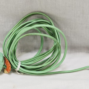 May include: A vintage green extension cord with a two-outlet cube and a plug. The cord is coiled, and a small tag is attached. The plug has two prongs and a grounding pin. The cord is made of a flexible, rubber-like material.