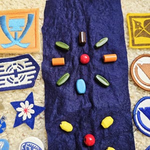 May include: An assortment of colorful patches and beads on a textured surface. The patches feature various geometric designs and colors, including orange, blue, yellow, and brown. The beads are in shades of red, green, yellow, and blue.