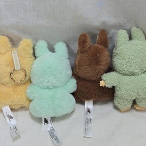 May include: A collection of six plush bunny toys in various colors: brown, yellow, light green, dark brown, green, and pink. Each bunny has a tag attached. The bunnies are standing upright, with their arms outstretched or at their sides.