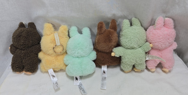 May include: A collection of six plush bunny toys in various colors: brown, yellow, light green, dark brown, green, and pink. Each bunny has a tag attached. The bunnies are standing upright, with their arms outstretched or at their sides.