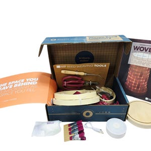 May include: A craft kit for making a woven reed luminary. The open box contains tools, reed strips, a wooden base, and a booklet. The kit includes pliers, a wooden handled tool, and a booklet with the text "Woven Reed Luminary".