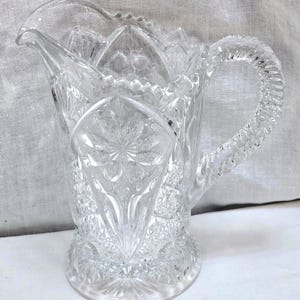 May include: Clear glass pitcher with a decorative, textured surface. The pitcher features a detailed pattern with a flared base, a curved handle, and a spout. The pitcher is designed for serving beverages.