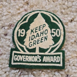May include: A vintage green and white felt Governor's Award patch. The patch features a tree design with the text "KEEP IDAHO GREEN" and the years "1950" and "19". The bottom of the patch reads "GOVERNOR'S AWARD".