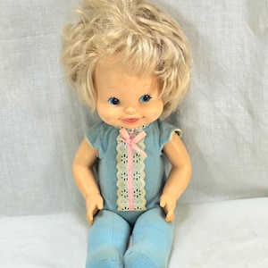 May include: A vintage doll with blonde hair, blue eyes, and a pink bow. She is wearing a blue and white outfit and is sitting on a white surface.