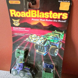 May include: A Matchbox Road Blasters toy car set featuring a green and black Road Razor vehicle with a snap-on armour feature. The set includes additional accessories, such as a green and black truck and a blue and black truck. The packaging features the text "Road Blasters" and "Power That Rules the Road!" along with the age recommendation "Ages 4 and up".