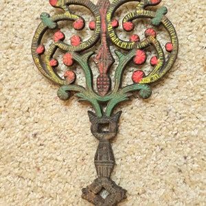 May include: A cast iron trivet with a decorative design featuring swirls, leaves, and flowers. The trivet is painted in shades of green, red, and yellow.