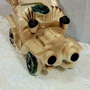 May include: A vintage ceramic planter shaped like a car. The car is beige with green wheels and brown accents. The planter has a scalloped top and a green and white floral design.