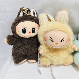 May include: Two plush dolls with smiling faces and furry costumes. One doll is brown with a bear-like hood, the other is yellow with bunny ears. Both have keychains attached.