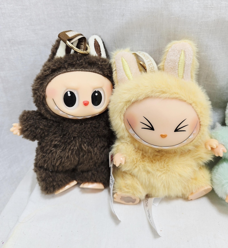 May include: Two plush dolls with smiling faces and furry costumes. One doll is brown with a bear-like hood, the other is yellow with bunny ears. Both have keychains attached.