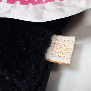 May include: Close-up of a black plush toy with a white and pink polka dot bow. A tag with the text "MINNIE MOUSE PRODUCT OF CONTENTS SYNTHETIC &copy; WALT DIS" is attached to the toy.
