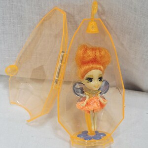 May include: A small fairy doll with bright orange hair, large dark eyes, and iridescent wings, wearing an orange skirt. The doll stands on a blue flower-shaped base inside a translucent orange, diamond-shaped plastic case with a top hanging hook.