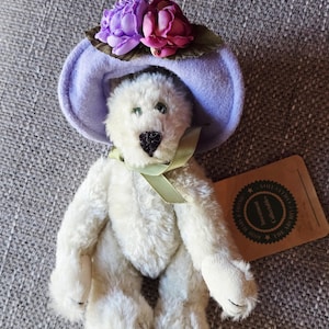 May include: A white teddy bear wearing a lavender hat adorned with purple and pink flowers. The bear has green eyes and a green ribbon. A brown tag is attached to the bear.