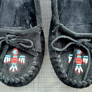 May include: A pair of black suede moccasins featuring a decorative beaded bird design on the vamp. The beadwork incorporates red, white, and light blue colors. Each shoe includes a black suede tie detail and visible whipstitch detailing along the edges.