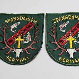 May include: Two green patches with the text "SPANGDAHLEM" and "GERMANY" in yellow. Each patch features a red banner with text, a yellow cross, two fish, and deer antlers.