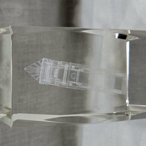 May include: A transparent, rectangular block with an etched architectural design inside. The design is of a building or structure with a pointed top. The block is clear, allowing the design to be viewed from all sides.