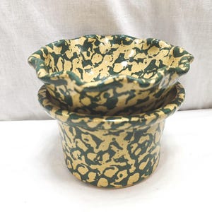 May include: Two-piece ceramic pottery set. The pieces are a light tan color with a dark green splatter pattern. The top piece is a small bowl with a ruffled edge. The bottom piece is a cylindrical container.