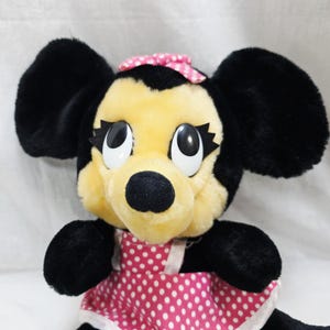 May include: A plush toy of a cartoon mouse with black ears, a yellow face, and a black nose. The mouse wears a pink dress with white polka dots and a matching bow. The toy has large, expressive eyes.