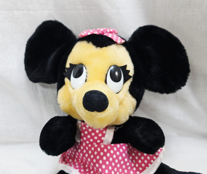 May include: A plush toy of a cartoon mouse with black ears, a yellow face, and a black nose. The mouse wears a pink dress with white polka dots and a matching bow. The toy has large, expressive eyes.