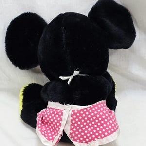 May include: A black plush toy with large, round ears and a pink polka-dot skirt. The toy has a white bow at the back of its neck and yellow accents on its feet. The toy is a classic character.
