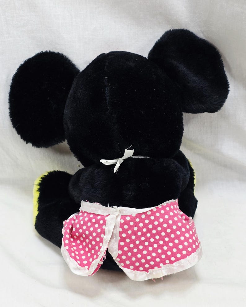 May include: A black plush toy with large, round ears and a pink polka-dot skirt. The toy has a white bow at the back of its neck and yellow accents on its feet. The toy is a classic character.