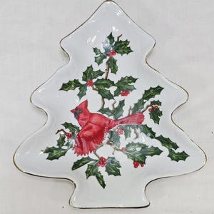 Op de afbeelding: Een witte keramische serveerschaal in de vorm van een kerstboom met een goudkleurige rand. Het midden is voorzien van een gedetailleerde illustratie van een felrode kardinaalvogel op groene hulsttakken met kleine rode bessen.