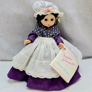 Puede incluir: Una muñeca Madame Alexander con un vestido morado y blanco y un gorro a juego. La muñeca tiene el pelo negro y una etiqueta que dice "Madame Alexander Miniature Showcase."