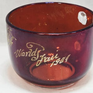 Puede incluir: Una taza de vidrio rojo oscuro con letras doradas que dicen "World's Fair 1904".