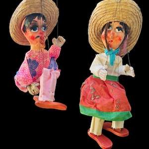 May include: Two vintage marionette puppets with straw hats and painted faces. One puppet wears a pink floral shirt and pink pants, the other wears a white shirt, red skirt, and a blue bow. Both have wooden shoes and are suspended by strings.