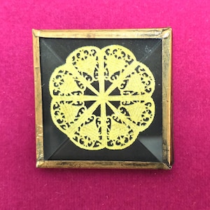 May include: Square artwork with a gold frame, showcasing a yellow, floral pattern on a black backdrop. The design is detailed and intricate. The artwork is set against a pink background.
