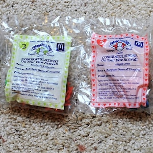 May include: Four sealed McDonald's Happy Meal toys featuring Cabbage Patch Kids. Each packet is a different color: blue, green, pink, and pink. Each packet has text that reads "Congratulations On Your New Arrival!" and the McDonald's logo.