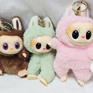 May include: Three plush keychains in brown, green, and pink. Each keychain features a cartoon character with large black eyes, a smiling mouth, and a fuzzy outfit with bunny ears. The keychains have gold-colored metal clasps.