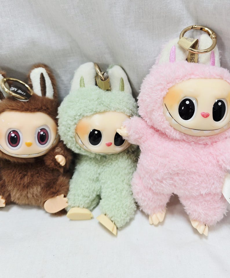 May include: Three plush keychains in brown, green, and pink. Each keychain features a cartoon character with large black eyes, a smiling mouth, and a fuzzy outfit with bunny ears. The keychains have gold-colored metal clasps.