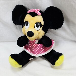 May include: A plush Minnie Mouse doll with black fur, large ears, and a yellow face. She wears a pink polka-dot dress and a matching bow. Her feet are yellow.