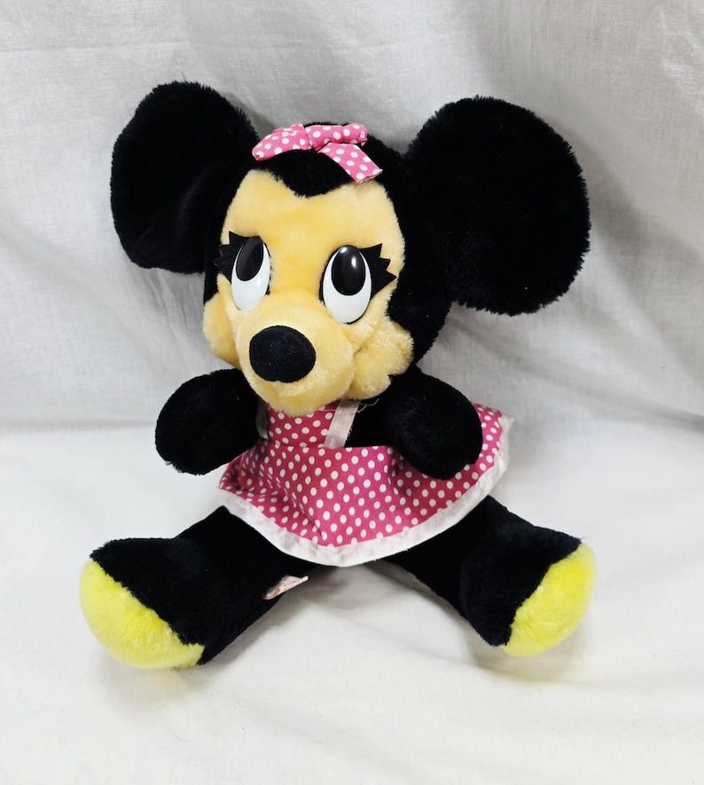 May include: A plush Minnie Mouse doll with black fur, large ears, and a yellow face. She wears a pink polka-dot dress and a matching bow. Her feet are yellow.