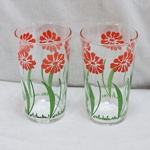 May include: Two clear drinking glasses with a floral design. The glasses feature red flowers with green stems and leaves. The flowers are painted on the outside of the glass, and the design is repeated around the glass.