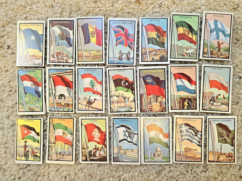 Op de afbeelding: Een verzameling van 20 kleine kaartjes, elk met de vlag van een ander land. De kaartjes zijn wit met een zwarte rand en een klein plaatje van een bezienswaardigheid of symbool uit het land. De vlaggen zijn allemaal in kleur en worden getoond op een witte paal.