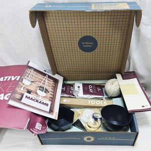 May include: An open craft kit box with macrame planter supplies. The box contains paper clay, clay tools, a ball of twine, and a black bowl. A booklet titled "Macrame Planters" is visible, along with a sticker that says "hi there, maker."