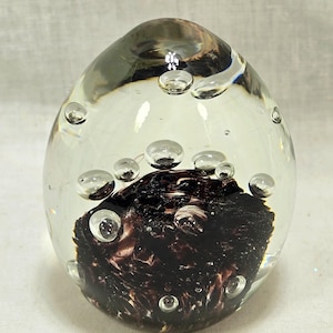 Może przedstawiać: Ciężarek do papieru ze szkła transparentnego z wirem ciemnofioletowego i czarnego szkła w środku. Ciężarek do papieru jest wypełniony małymi pęcherzykami powietrza.