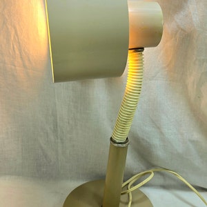 May include: A beige desk lamp with a cylindrical base and a flexible, ribbed neck. The lamp has a rectangular shade and a cord. The light is on, illuminating the shade's interior. The lamp is made of metal.