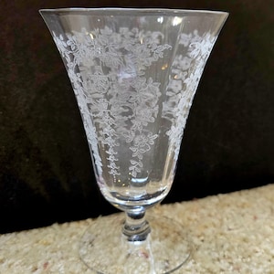 Könnte beinhalten: Ein klares Glas mit Stiel und einem geätzten Blumendesign. Das Glas hat eine ausgestellte Form und einen runden Fuß. Das florale Muster bedeckt das gesamte Glas und erzeugt ein zartes und elegantes Aussehen. Das Glas wird wahrscheinlich zum Servieren von Getränken verwendet.